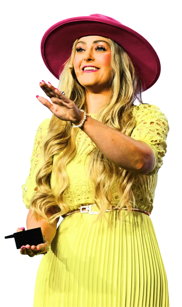 Stefanie Couch speaking on stage in a yellow dress and pink hat, one hand raised