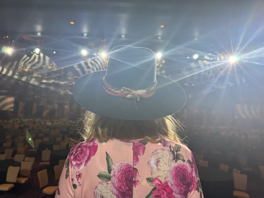Stefanie Couch, seen from behind in her hat, facing the empty Employee Owned 2025 ballroom as the stage lights flare