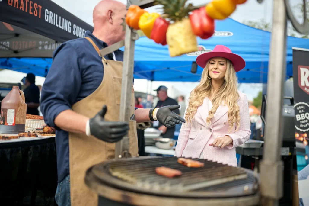A grill chef turns sausages under a skewer of peppers and pineapple as Stefanie laughs beside his grill