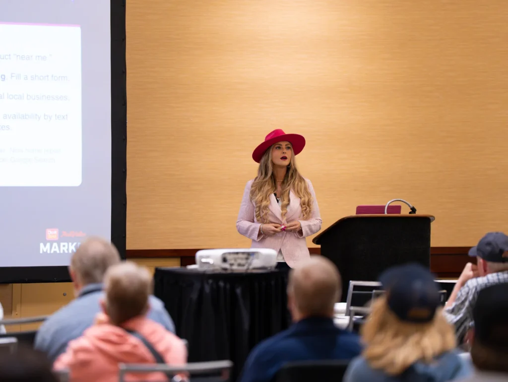 Hands together at the front of the room, Stefanie leads her Millwork Academy session beside the lectern