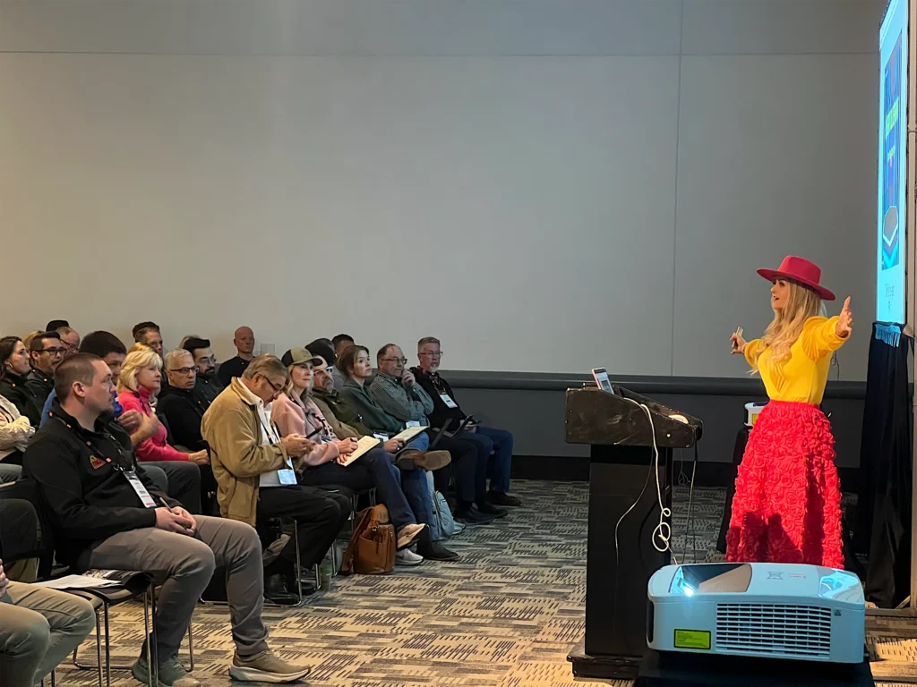 Arms out beside the lectern, Stefanie speaks to a room of attendees taking notes at Spring Market 2026