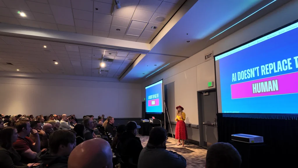 A large audience watches Stefanie Couch present a slide that reads AI Doesn't Replace the Human
