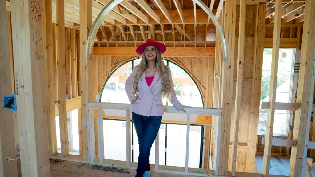 Stefanie Couch in a pink hat and light pink blazer leaning on a railing inside a house under construction