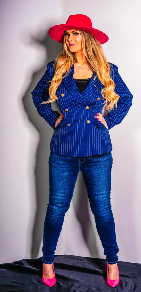 Stefanie Couch standing with her hands on her hips in a navy pinstripe blazer, jeans and pink hat